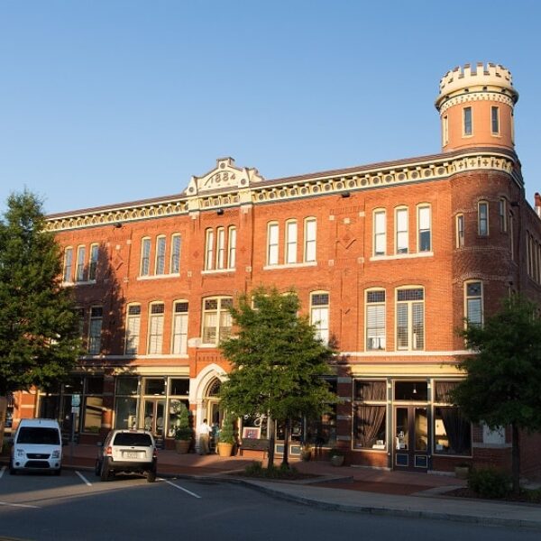 A historic, three-story brick building with arched windows and a distinctive turret on the corner. The building is fronted by trees and several parked vehicles, including a white van and a yellow truck, on a sunny day with a clear blue sky.