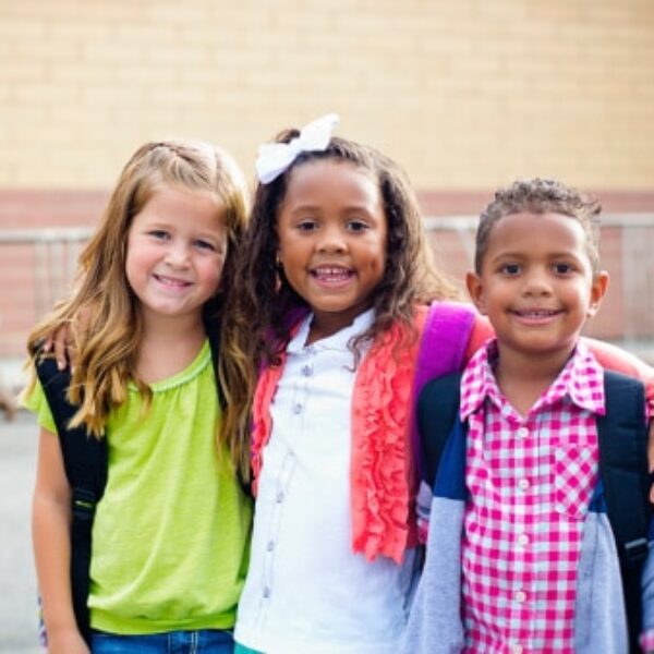 Three children stand side by side, smiling at the camera. Each child has a backpack on, indicating they are ready for school. They are outdoors, with a building and metal bike racks in the background. The child in the center has their arms around the other two.