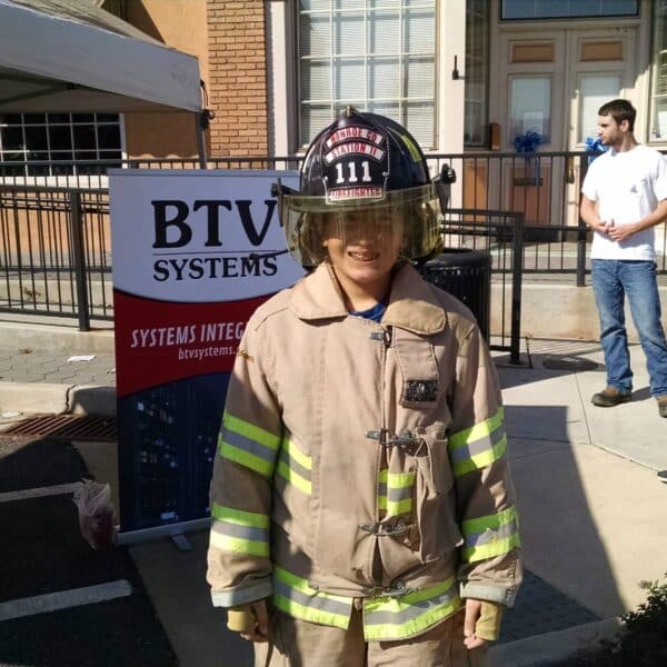 A child is smiling and wearing a firefighter's helmet and jacket. Behind the child is a booth with a banner that reads 