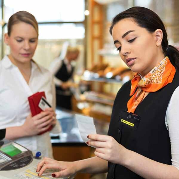 Two women are at a counter in what appears to be a bakery or café. One is a customer holding a wallet, and the other, likely an employee, is wearing a uniform and looking at a receipt or paper. The background shows baked goods on display.