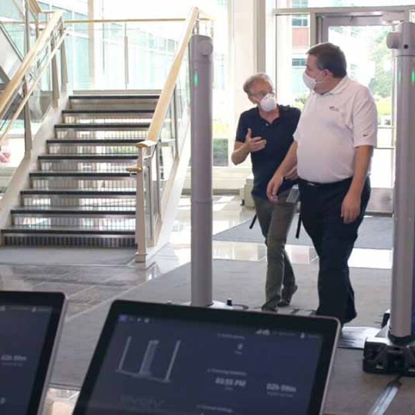 Two people in masks walk past security scanners inside a modern building. One person is gesturing while the other listens. In the foreground, computer screens display data. A staircase is visible in the background leading to an upper level.