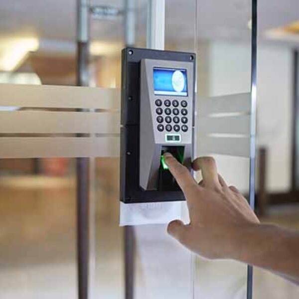 A person is using a biometric fingerprint scanner mounted on a glass door. The device also features a numeric keypad and a small display screen. The background shows an indoor environment with blurred furnishings and lights.