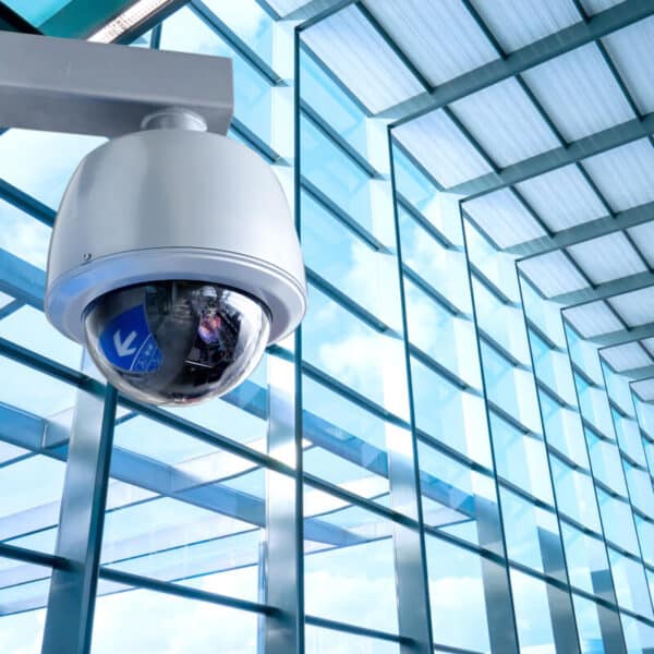 A security camera mounted on a wall inside a modern building with large glass windows and a high ceiling. The camera, enclosed in a white dome, points towards a bright, expansive area.