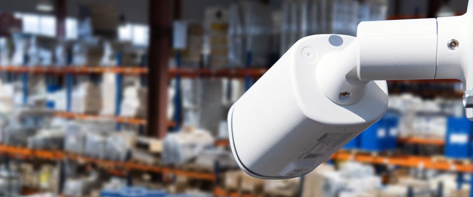 Close-up of a security camera indoors, with a blurred background showing shelves stocked with various items, suggesting a warehouse or storage facility.