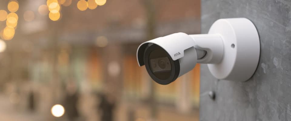 A white security camera is mounted on a gray wall, facing an outdoor area with blurred lighting and people in the background. The camera has a sleek design and possibly features night vision capabilities.