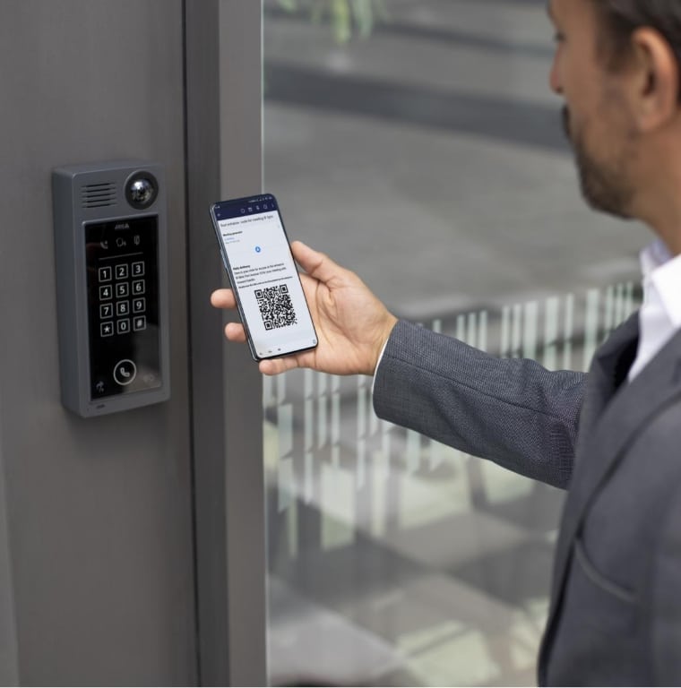 A person in a suit holds a smartphone displaying a QR code towards a keypad access control system mounted on a wall. The system includes a numeric keypad, camera, and speaker for secure entry. The background features a modern building interior with glass and metal elements.