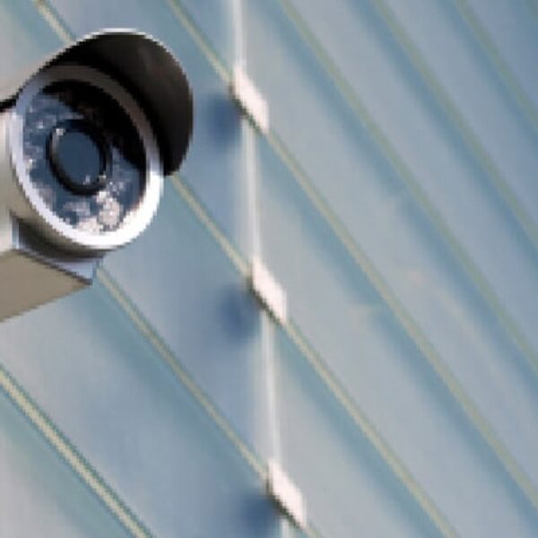 A silver-colored surveillance camera mounted on an exterior wall of a building, angled slightly downward. The wall features a repeating pattern of diagonal lines, creating a textured backdrop. The camera lens is prominently visible within its cylindrical housing.