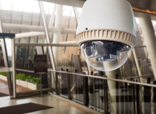 A white security camera dome is mounted on the ceiling of a modern, glass-walled building with plants and railings in the background. The camera lens is visible inside the transparent dome, and the setting appears to be a public or commercial space.