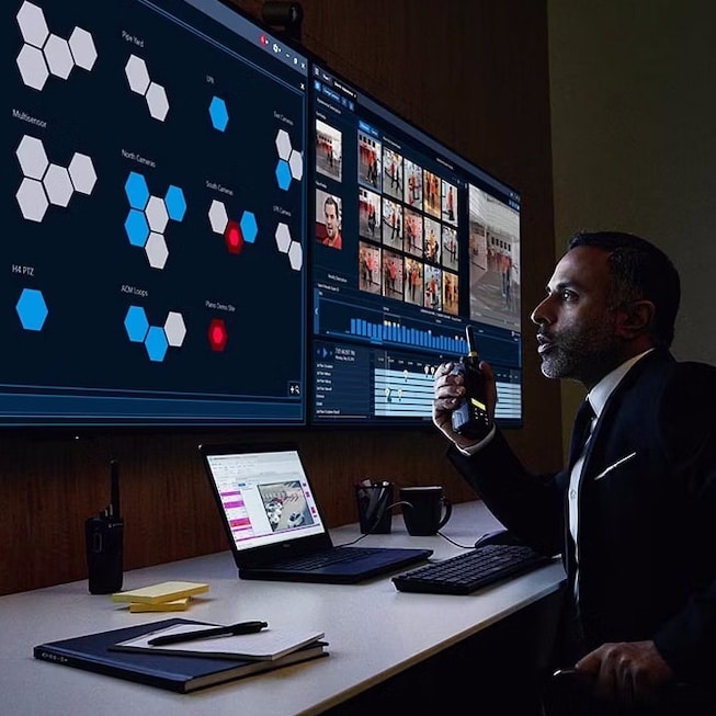 A man in a suit sits at a desk with multiple monitors. One screen shows hexagonal data points, another displays surveillance footage. He holds a walkie-talkie and appears focused. The desk has a laptop, notepads, and two cups. The setting looks like a control room.