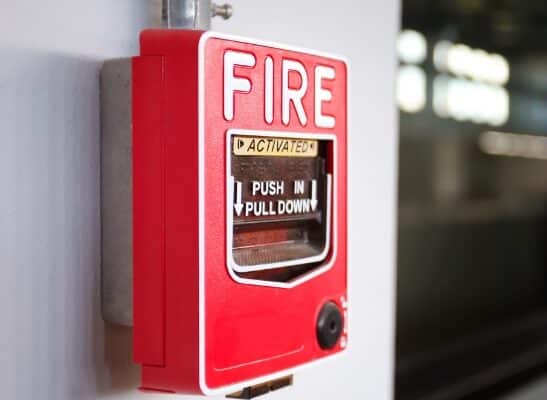 A red fire alarm is mounted on a white wall with a metallic pipe connected at the top. The alarm reads "FIRE" in large white letters, with instructions to "PUSH IN PULL DOWN" below. A small black circular element is located at the bottom right.