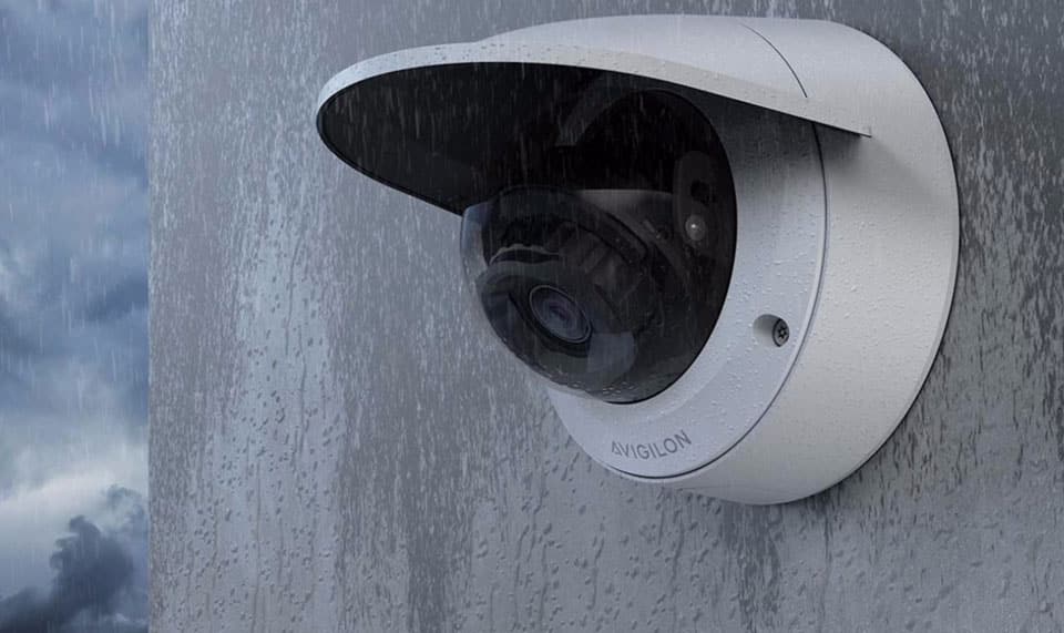 A security camera mounted on a wet wall in rainy conditions. The camera has a dome shape with a protective cover, shielding it from the rain. The sky is overcast, and rain droplets are visible on the camera and wall.