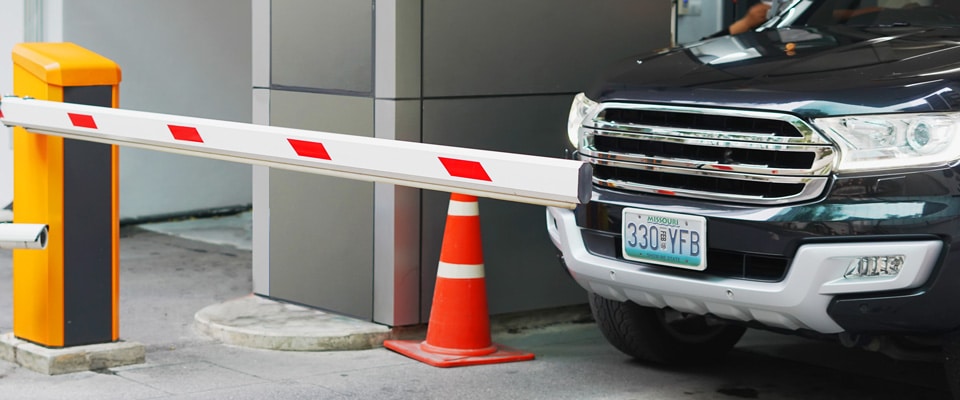 A black SUV is stopped at a parking gate with a white and red barrier arm blocking its path. An orange and white traffic cone stands near the barrier system, which includes a yellow control box adjacent to a grey building. The SUV has a visible license plate.