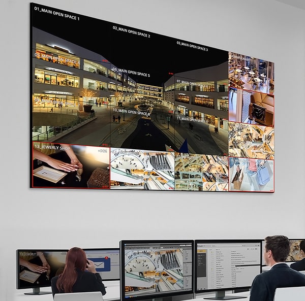 A security control room with two individuals monitoring multiple surveillance camera feeds displayed on a large screen. The screen shows various angles of a retail space, including a jewelry store and different open spaces. Smaller screens are visible below.