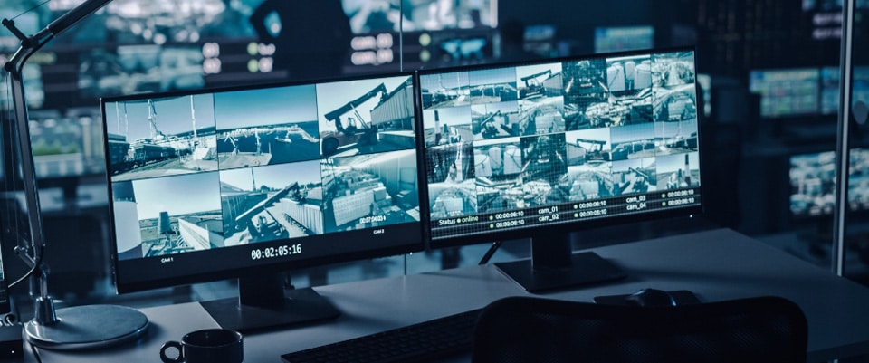 A workspace featuring two monitors displaying multiple CCTV footage screens of outdoor locations. The desk holds a keyboard, a lamp, and a cup, with more surveillance screens visible in the background. The atmosphere is dim, indicating a control or monitoring room.