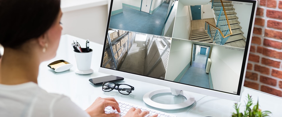 A person sitting at a desk, monitoring security footage on a computer with a white keyboard. The screen displays four different surveillance camera views of an indoor area, including hallways and a staircase. Nearby are glasses, a cup, a mobile phone, and a notebook.