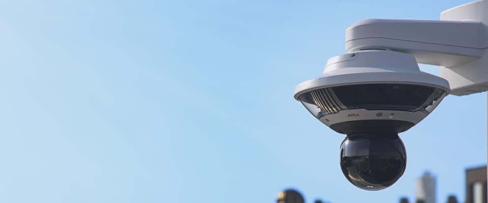 A white, dome-shaped security camera is mounted on an outdoor wall, angled slightly downward. The camera is set against a clear blue sky, with the tops of buildings blurred in the background.