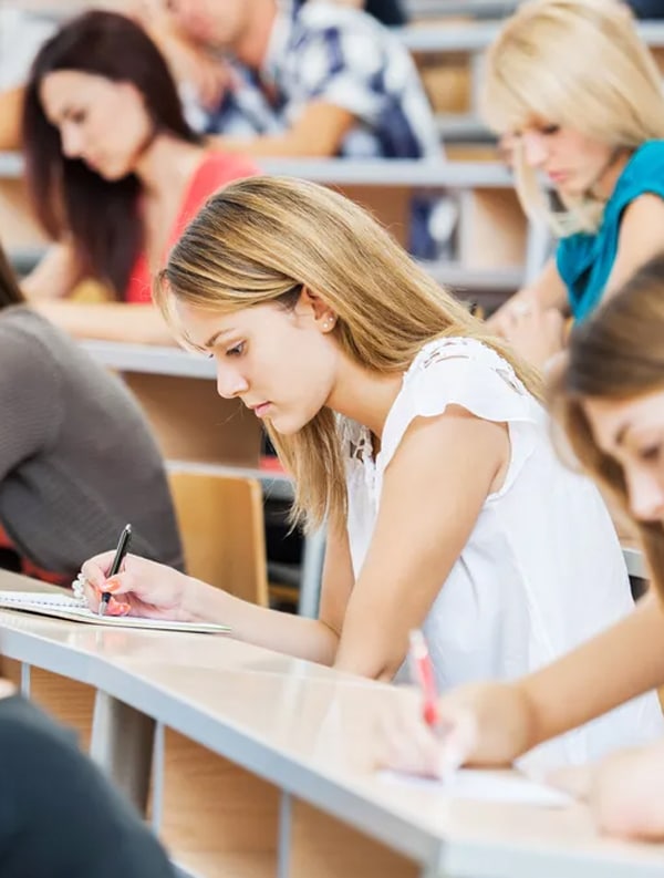 A classroom setting with several students sitting at desks, focusing intently on writing in notebooks. The foreground features a young woman with long blonde hair, wearing a white top, deeply absorbed in her work. Other students are visible in the background.