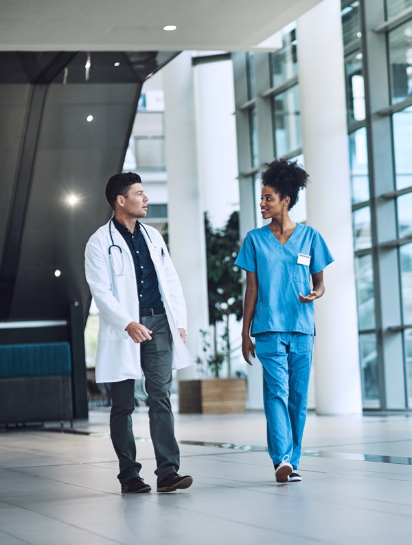 A doctor in a white coat and a nurse in blue scrubs walk and converse in a spacious, modern hospital lobby with large windows. The doctor holds a stethoscope, and the nurse gestures with one hand while holding medical files in the other.