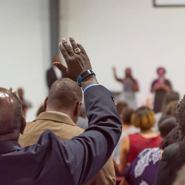 A person in a suit raises their hand in a room with other people. The scene appears to be indoors, possibly at a meeting or an event, with blurred figures in the background suggesting action or dialogue taking place in front of an audience.