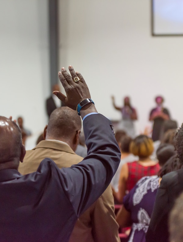 A person in a suit raises their hand in a room with other people. The scene appears to be indoors, possibly at a meeting or an event, with blurred figures in the background suggesting action or dialogue taking place in front of an audience.